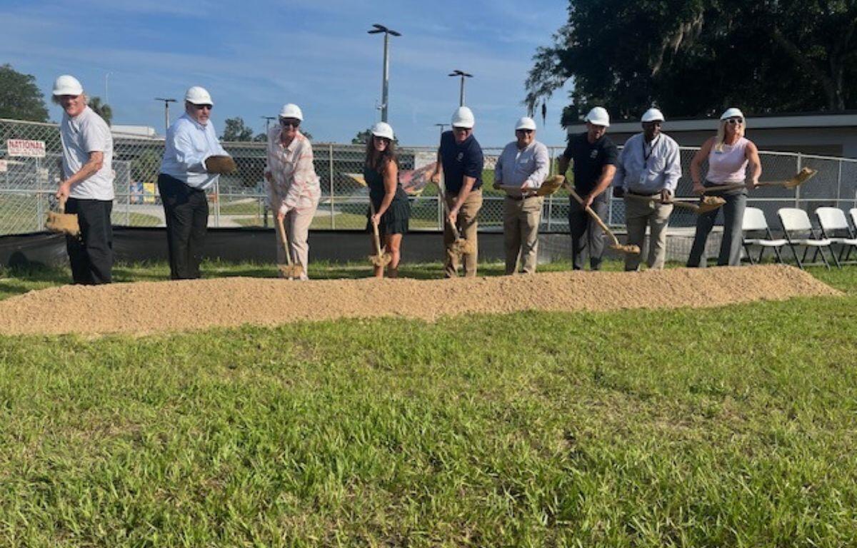 A group of people wearing hardhats, with shovel that have dirt on the blade, with lighting fixtures in the background, dirt directly in front of them, and a grass field in the foreground.