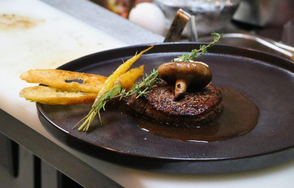 Seared meat with rich sauce, garnished with vegetables and crispy plantain chips on a dark plate.
