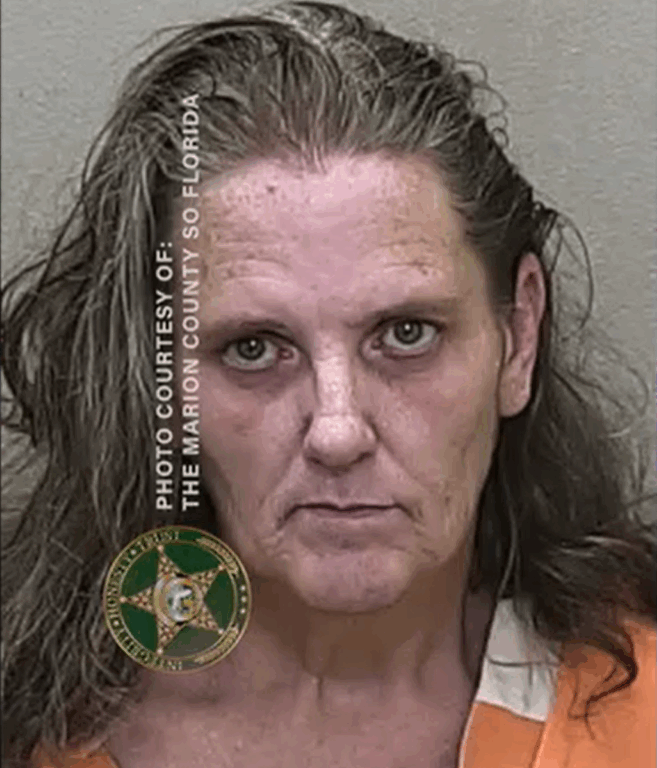 Mugshot of a woman with long, wavy hair wearing an orange jail uniform, facing the camera with a neutral expression.