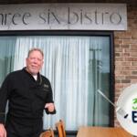 A man in a black chef's uniform standing in front of a brick-front underneath a sign that reads, "Three Six Bistro".