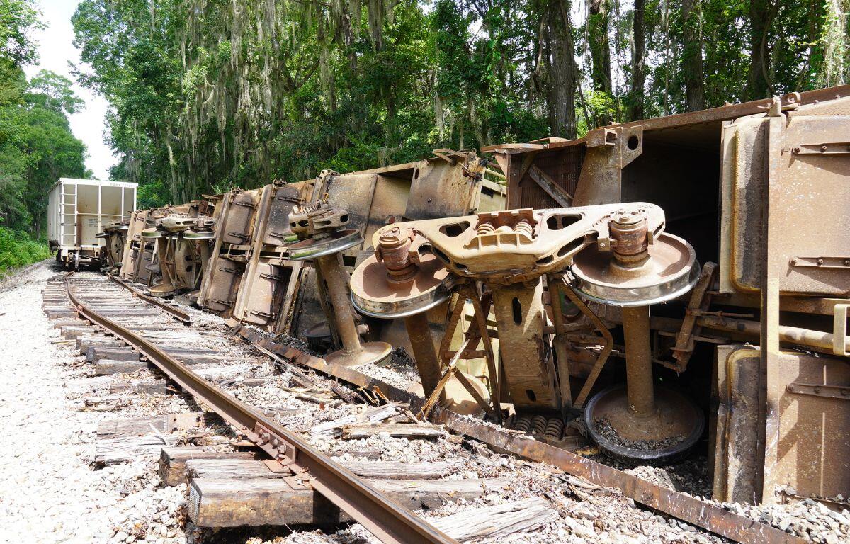 The left side, a railroad track, of the to the right, derailed train cars, to the extreme right and in the background tree lines.