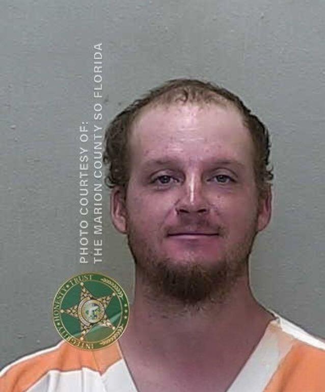 Mugshot of a man with short light brown hair and a light beard, wearing an orange-and-white striped prison uniform.