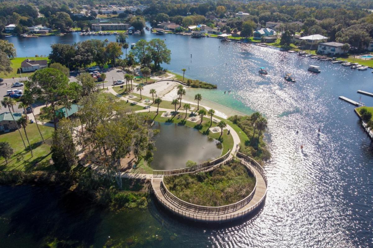 Aerial view of a scenic waterfront park featuring a curved boardwalk over a pond, sandy beach area, palm trees, walking paths, and several boats floating on a calm, sunlit river surrounded by residential homes and greenery.