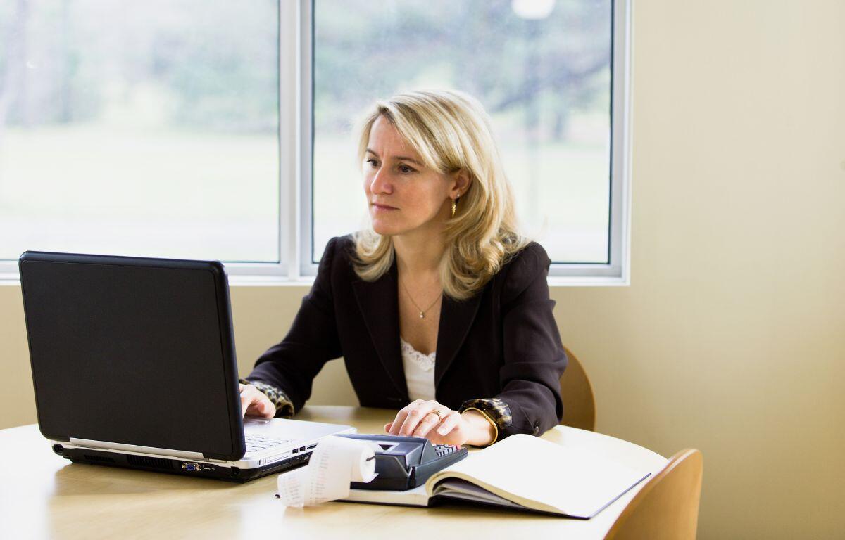 A professional woman in a black blazer works at a laptop in a bright office, with an open notebook and calculator on the desk, focused on her financial documents.