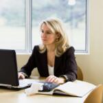 A professional woman in a black blazer works at a laptop in a bright office, with an open notebook and calculator on the desk, focused on her financial documents.