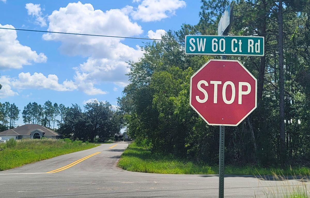 A red stop sign is positioned at the corner of a rural intersection, with a green street sign above it reading "SW 60 Ct Rd." The road curves to the left, bordered by grass and trees. A house is visible in the background under a blue sky with scattered white clouds.