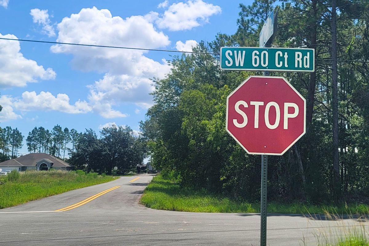 A red stop sign is positioned at the corner of a rural intersection, with a green street sign above it reading "SW 60 Ct Rd." The road curves to the left, bordered by grass and trees. A house is visible in the background under a blue sky with scattered white clouds.