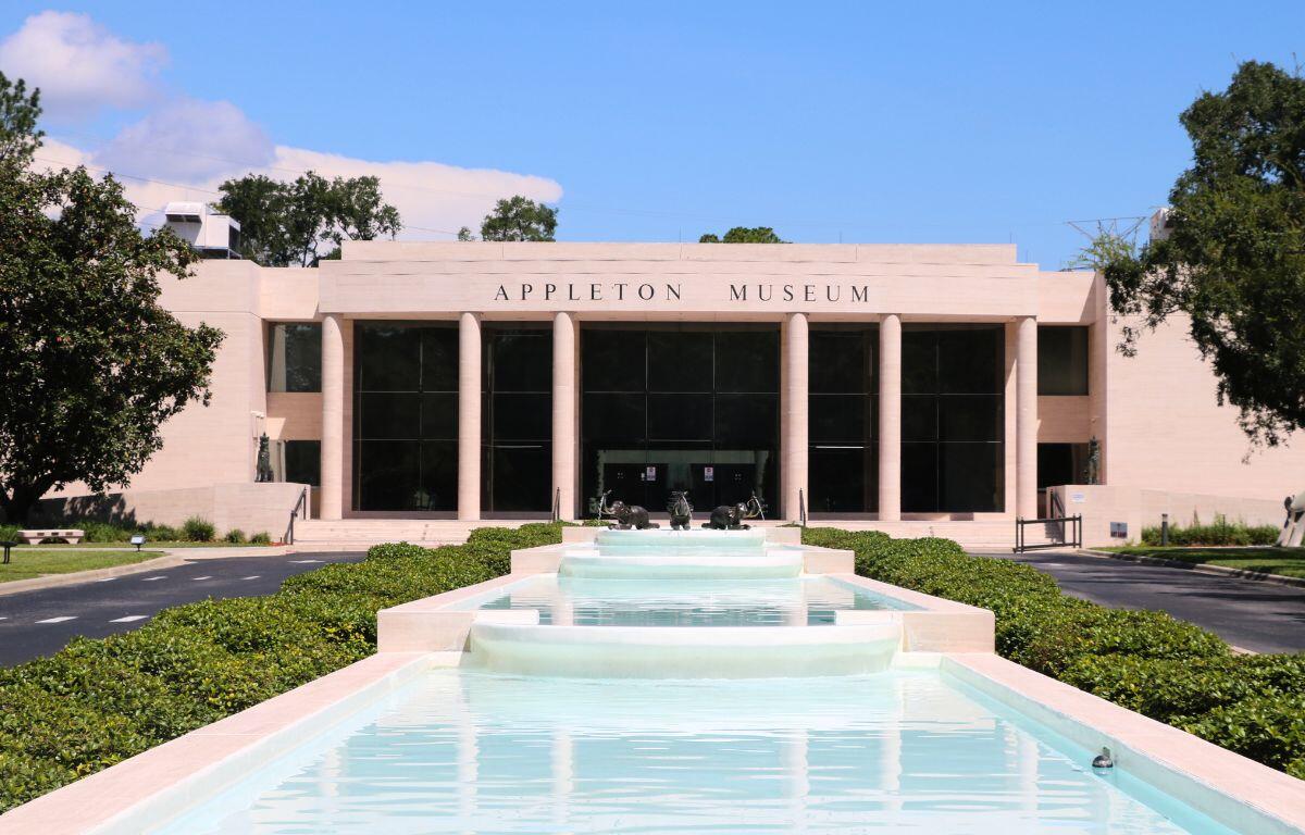 Front view of the Appleton Museum of Art, featuring a grand modern facade with tall columns and large dark-tinted glass windows. A cascading water fountain runs down the center of the entrance walkway, surrounded by neatly trimmed greenery and a circular driveway. Three decorative animal sculptures are placed in the middle of the fountain near the museum entrance, and the sky is clear and blue.