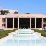 Front view of the Appleton Museum of Art, featuring a grand modern facade with tall columns and large dark-tinted glass windows. A cascading water fountain runs down the center of the entrance walkway, surrounded by neatly trimmed greenery and a circular driveway. Three decorative animal sculptures are placed in the middle of the fountain near the museum entrance, and the sky is clear and blue.