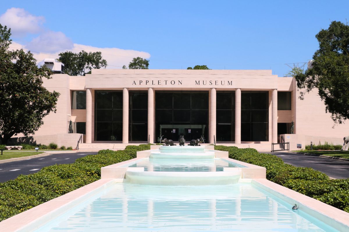 Front view of the Appleton Museum of Art, featuring a grand modern facade with tall columns and large dark-tinted glass windows. A cascading water fountain runs down the center of the entrance walkway, surrounded by neatly trimmed greenery and a circular driveway. Three decorative animal sculptures are placed in the middle of the fountain near the museum entrance, and the sky is clear and blue.