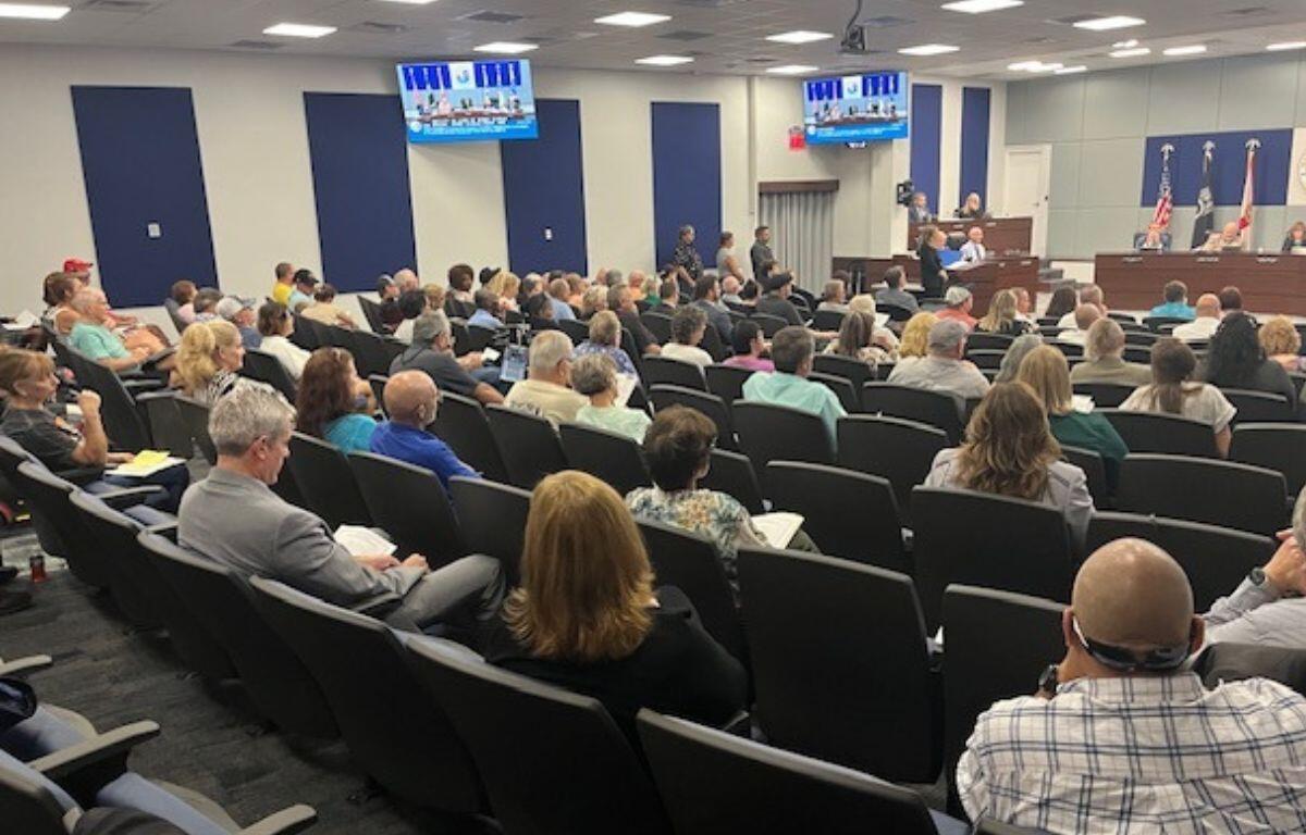 An auditorium with a sea of people sitting in seats, two overhead screens against the left and left-center wall, with someone speaking at a podium on the left, and people sitting behind a tiered dais, with flags and insignia in the background.