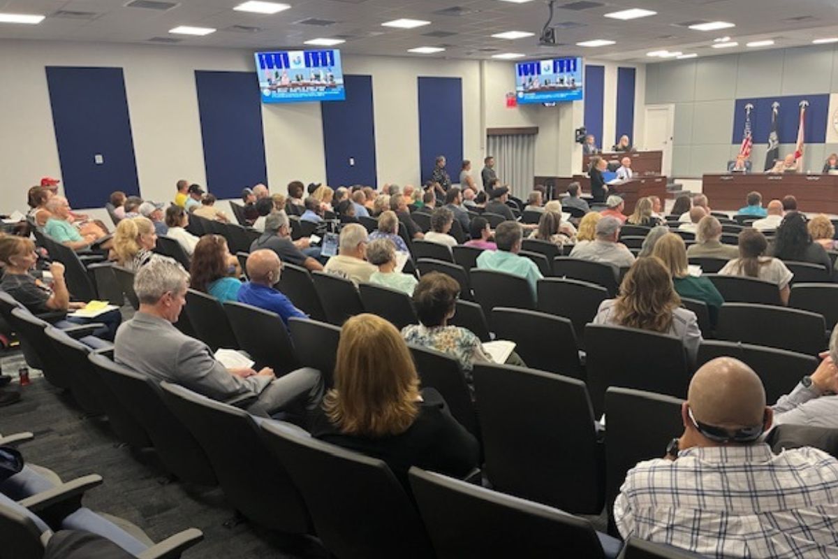 An auditorium with a sea of people sitting in seats, two overhead screens against the left and left-center wall, with someone speaking at a podium on the left, and people sitting behind a tiered dais, with flags and insignia in the background.