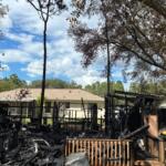 Charred remains of a shed in the foreground, with trees to the right, center and left, a dwelling in the background and another to the right.