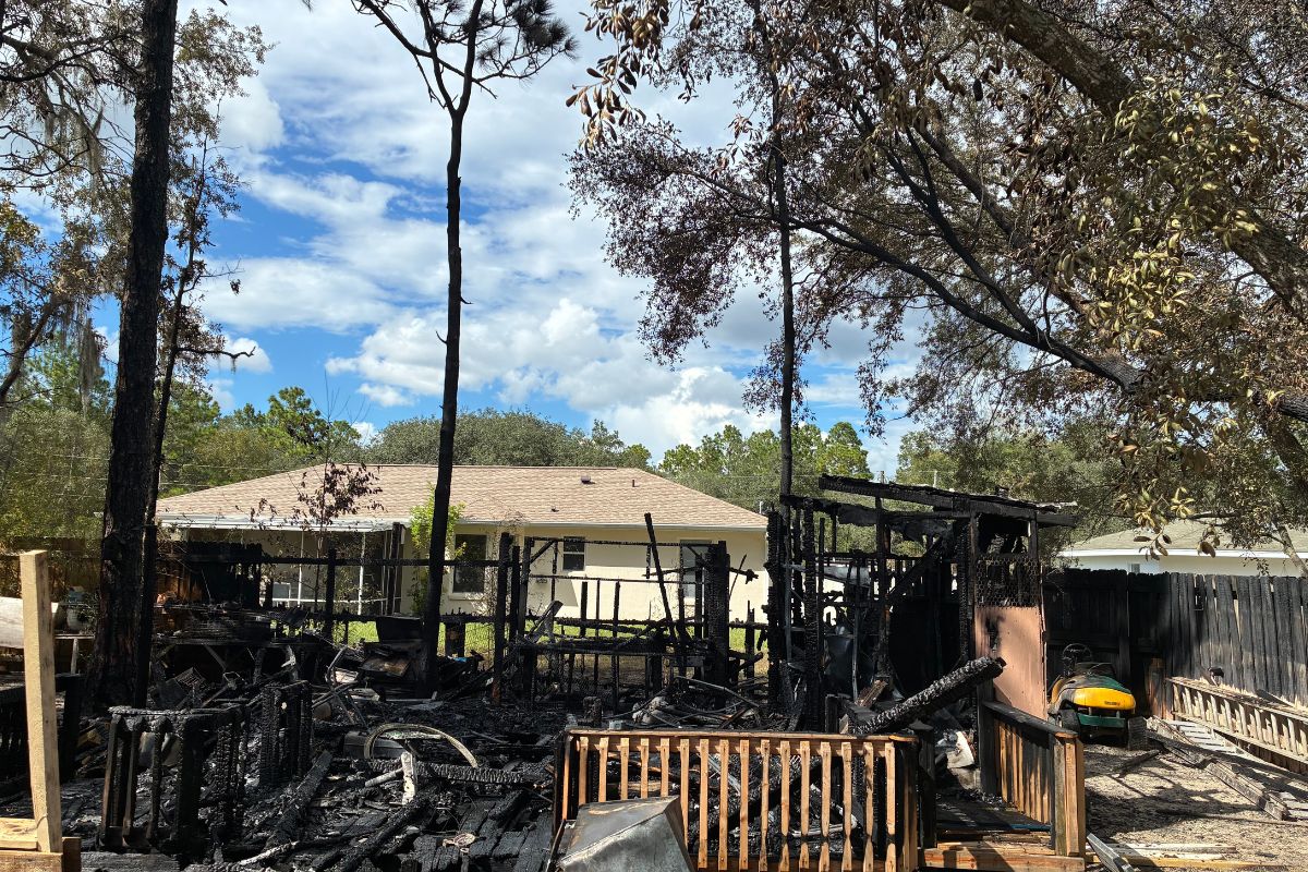 Charred remains of a shed in the foreground, with trees to the right, center and left, a dwelling in the background and another to the right.