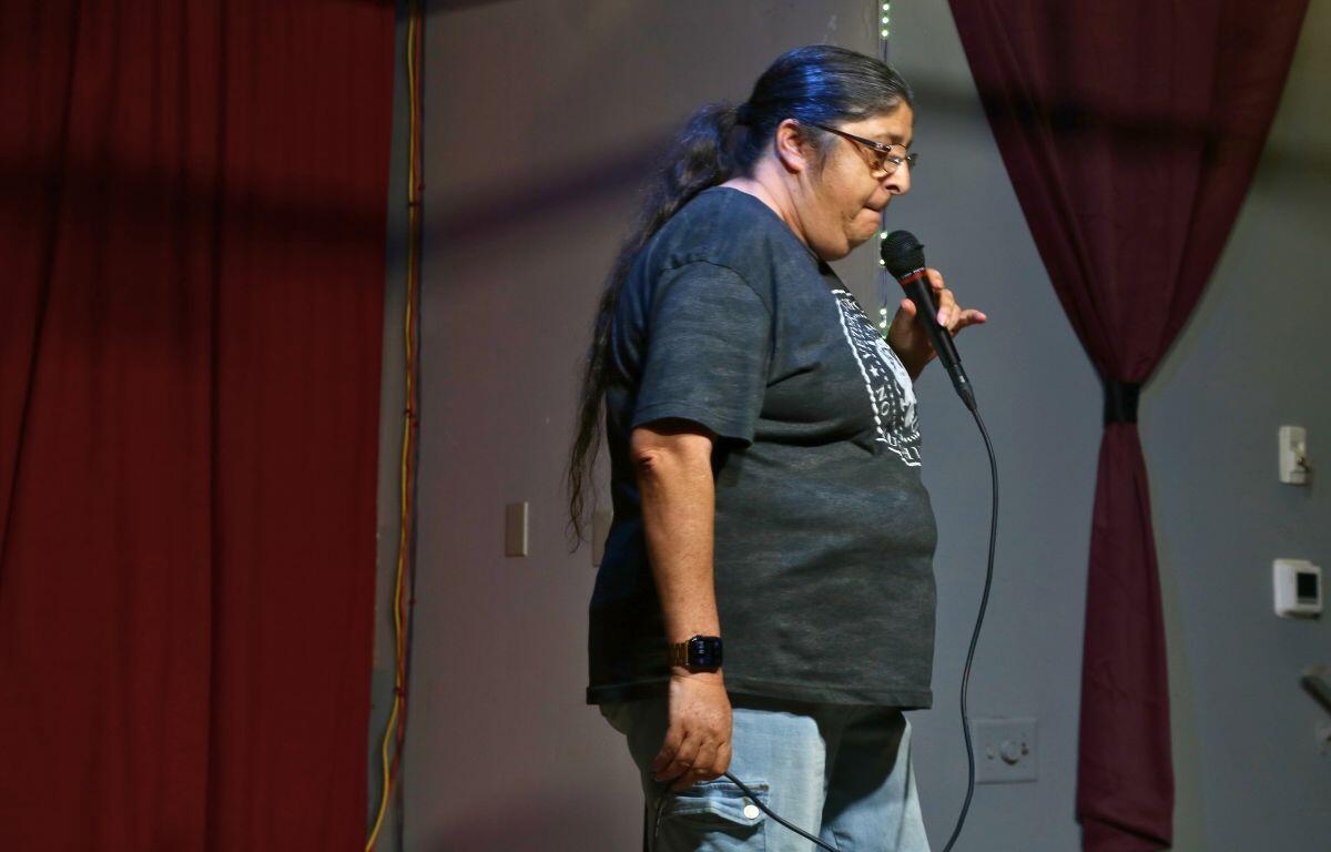 A comedian stands onstage delivering jokes, speaking into the microphone in front of red curtains.