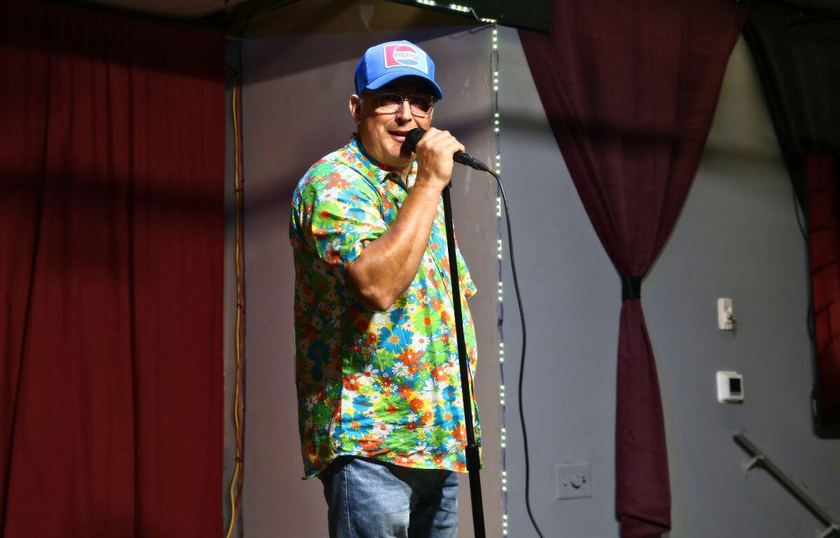 A comedian in casual attire performs onstage with microphone in hand.