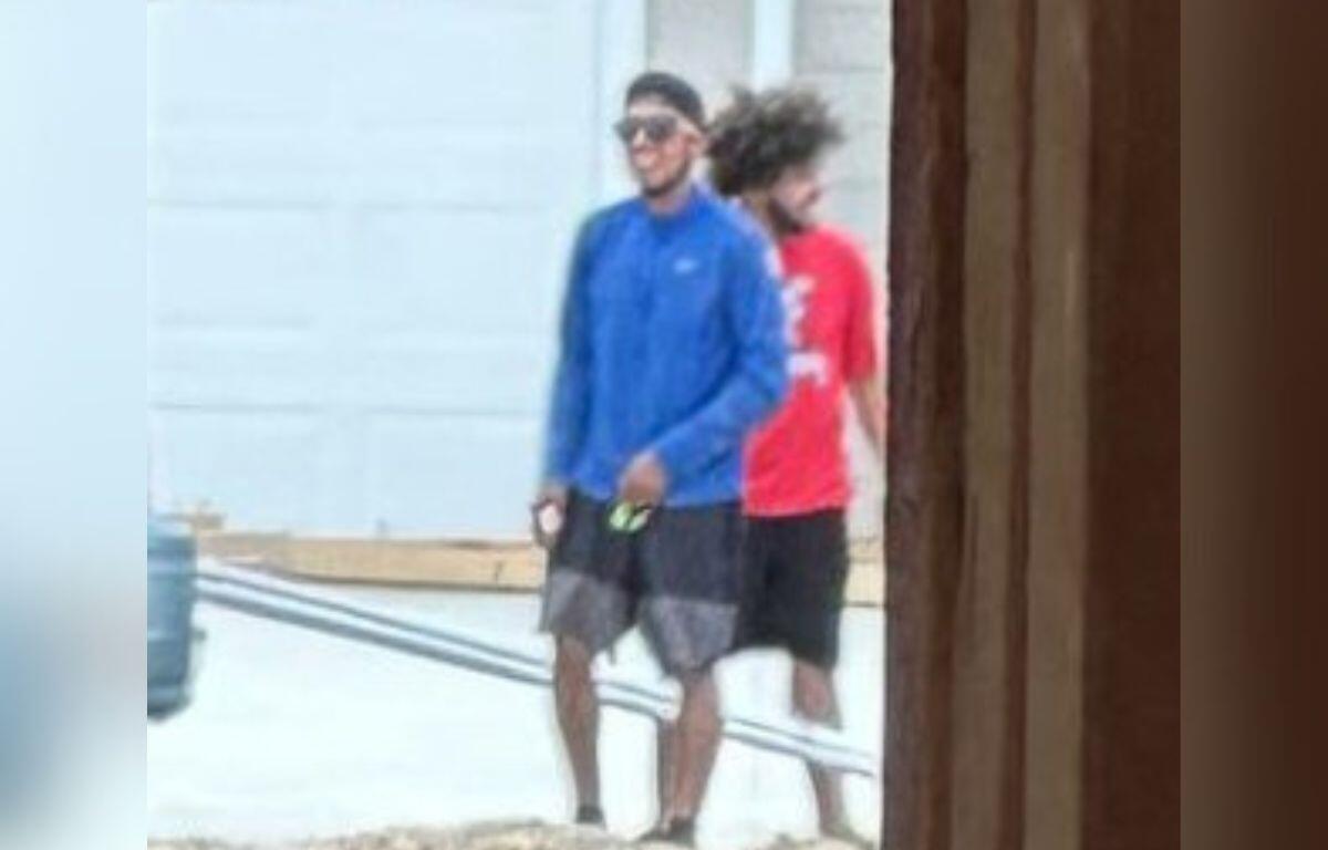 Two men walking outside, one wearing a blue shirt and the other in red, near a residential area.