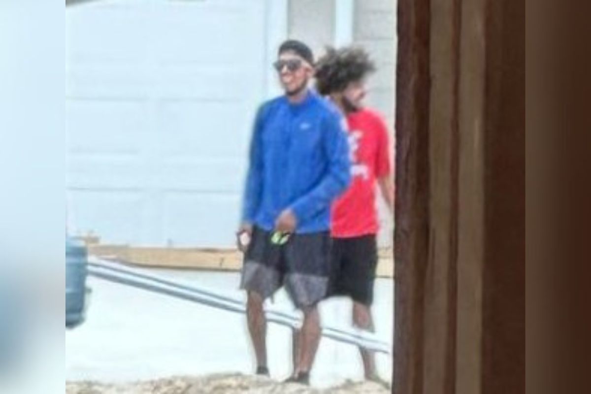 Two men walking outside, one wearing a blue shirt and the other in red, near a residential area.