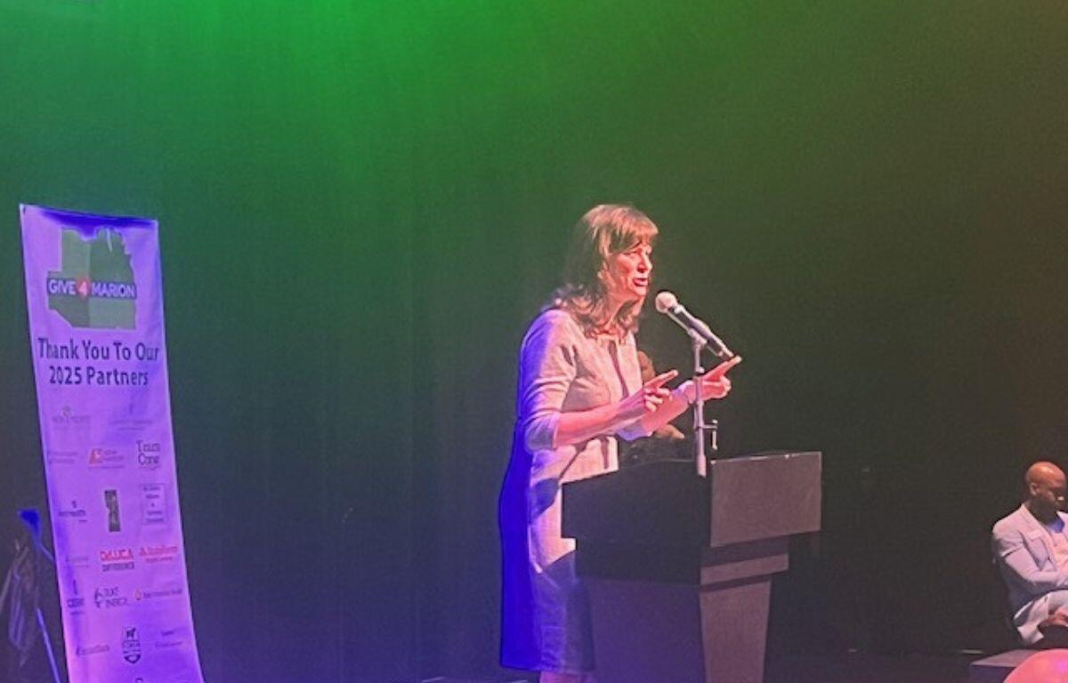 A well-dressed woman standing behind a podium with a microphone, with a background of black curtains, with a banner behind her to the left.