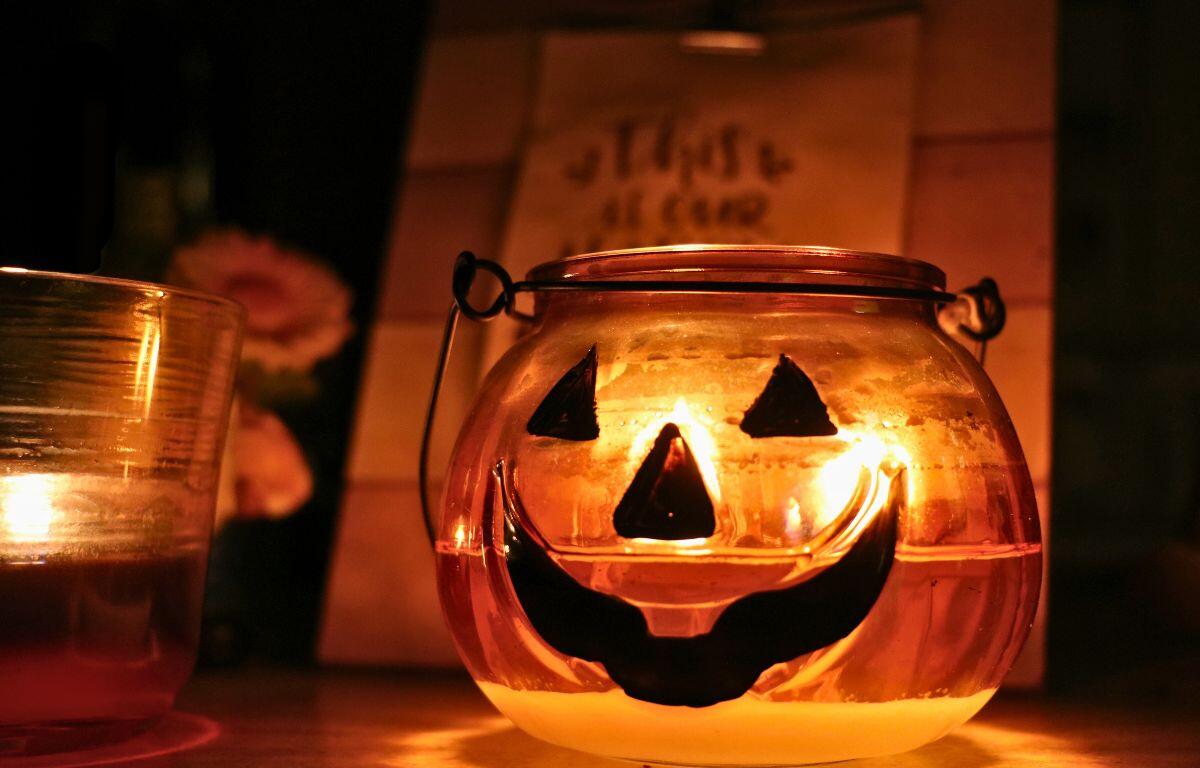 A pumpkin candle holder with a face painted to look like a jack-o-lantern with another red candle to the left of it. In the backgroud is a blurred wooden sign and a small gnome with a flower.