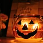 A pumpkin candle holder with a face painted to look like a jack-o-lantern with another red candle to the left of it. In the backgroud is a blurred wooden sign and a small gnome with a flower.