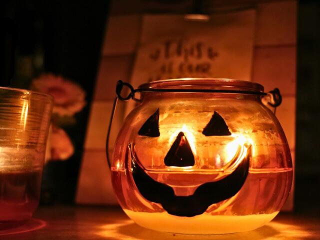 A pumpkin candle holder with a face painted to look like a jack-o-lantern with another red candle to the left of it. In the backgroud is a blurred wooden sign and a small gnome with a flower.