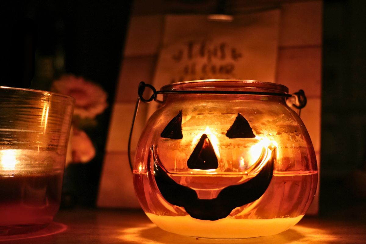 A pumpkin candle holder with a face painted to look like a jack-o-lantern with another red candle to the left of it. In the backgroud is a blurred wooden sign and a small gnome with a flower.