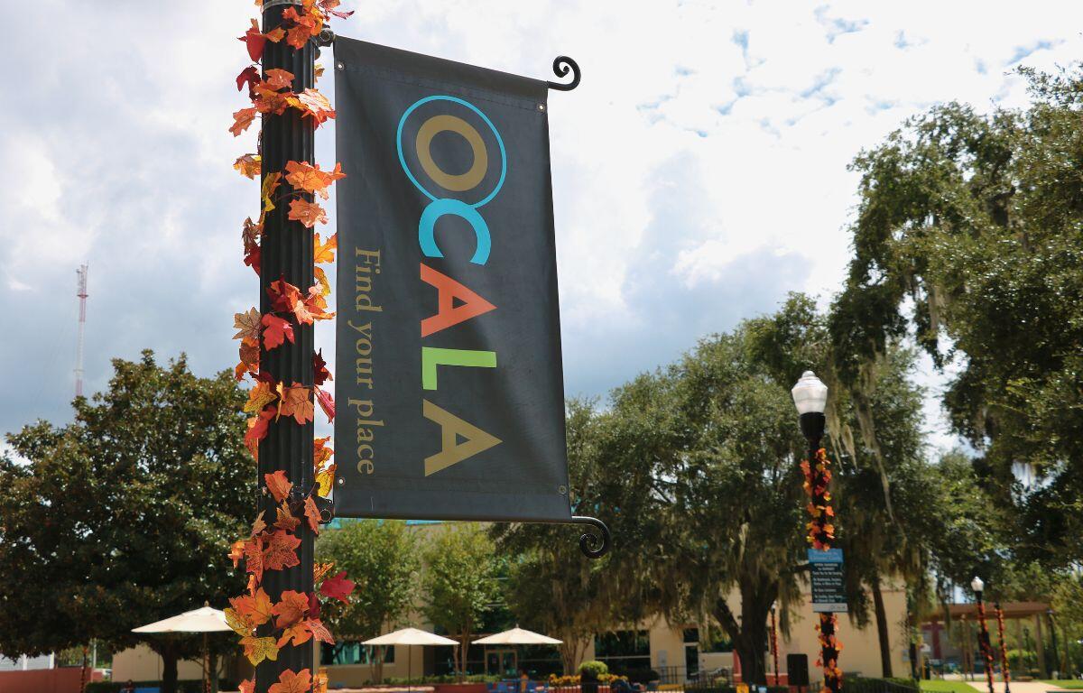 In the foreground is a light pole with autumn-colored leave and a sideways sign reading, "Ocala. Find your place." In the background are trees, buildings and multiple poles with similar leaves and signs reading rules. There are also tables with umbrellas and walking paths for public use.
