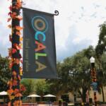 In the foreground is a light pole with autumn-colored leave and a sideways sign reading, "Ocala. Find your place." In the background are trees, buildings and multiple poles with similar leaves and signs reading rules. There are also tables with umbrellas and walking paths for public use.