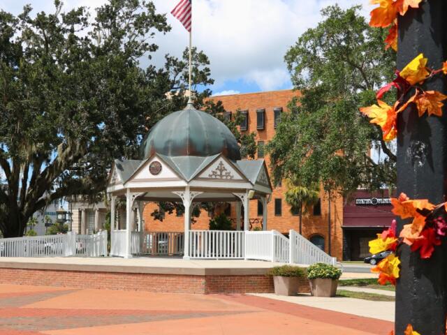 In the foreground is a blurred black light pole with autumn-colored leaves hanging around it. In the background is a white gazebo with an American flag standing on top of it. Further back are restaurants and shops lining a city street.