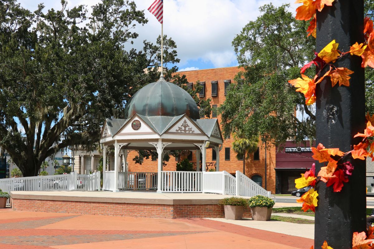 In the foreground is a blurred black light pole with autumn-colored leaves hanging around it. In the background is a white gazebo with an American flag standing on top of it. Further back are restaurants and shops lining a city street.