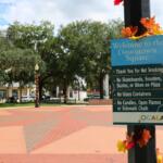 In the foreground is a black light pole with autumn-colored leaves hanging from it and a sign that reads the rules and for Downtown Square Ocala. In the background is a large, open space with a view of a variety of shops and restaurants.