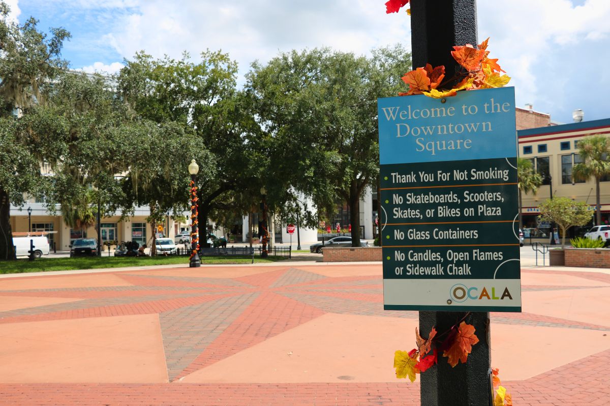 In the foreground is a black light pole with autumn-colored leaves hanging from it and a sign that reads the rules and for Downtown Square Ocala. In the background is a large, open space with a view of a variety of shops and restaurants.