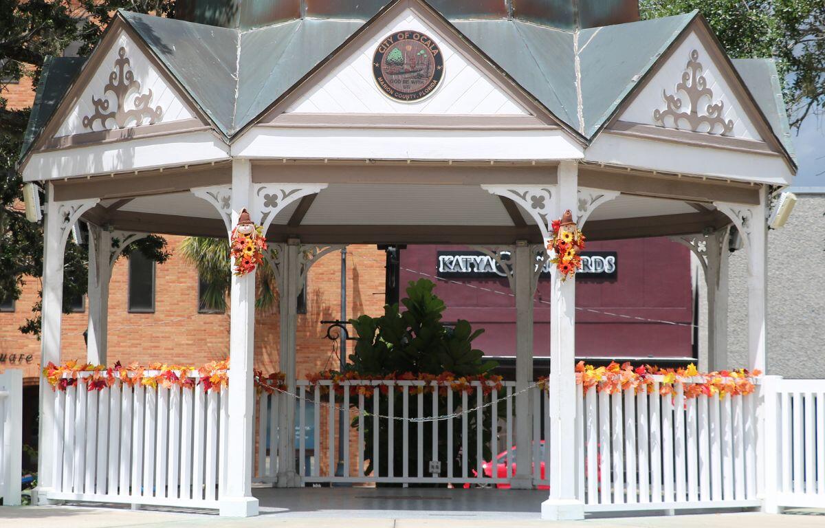 A white gazebo with a green roof and circular medallion at the top reading, "City of Ocala. Marion County, Florida" sits in the foreground with brick-buildings and shops in the background. The gazebo has fall-colored leaves surrounding it's railing and fall-colored flowers on either side of its entrance with little scarecrows on top of them.