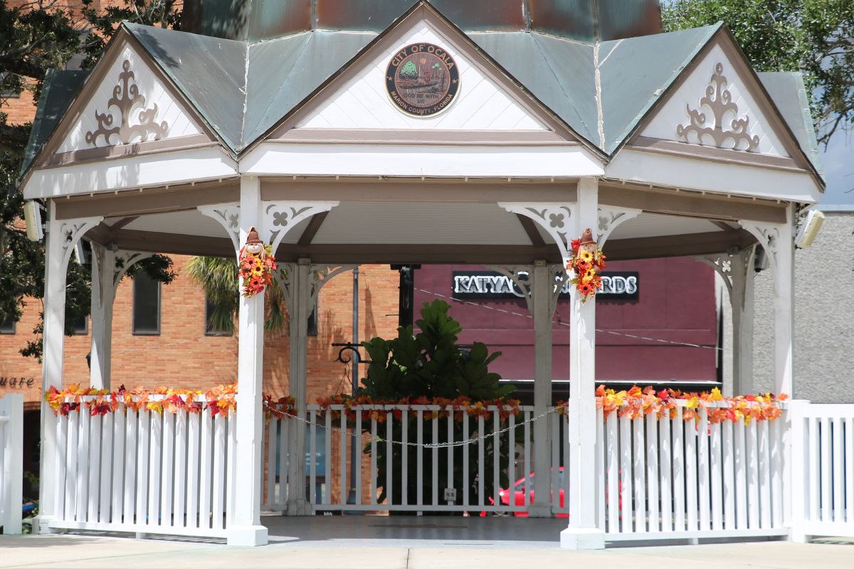 A white gazebo with a green roof and circular medallion at the top reading, "City of Ocala. Marion County, Florida" sits in the foreground with brick-buildings and shops in the background. The gazebo has fall-colored leaves surrounding it's railing and fall-colored flowers on either side of its entrance with little scarecrows on top of them.