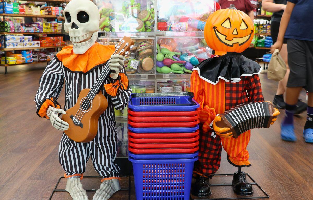 On the left is a skeleton in a black, white and orange clown outfit with a miniature guitar. On the right is a pumpkin in an orange, red, black and white clown outfit holding an accordion. In the center are red and blue shopping baskets with various candy and toy items on shelf in the background Grandpa Joe's Candy Shop.