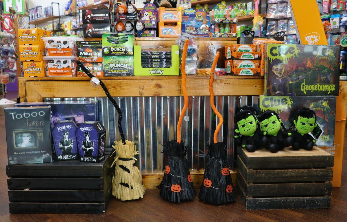 A variety of decor and items including witch brooms, Wednesday toys, Halloween-themed candy, Jason toys and more are on display in front of and on top of a stand in Grandpa Joe's Candy Shop.