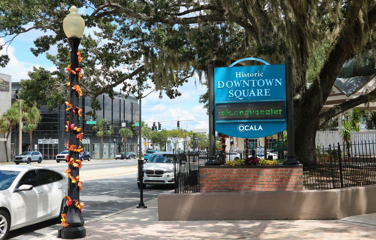 On the left is a light pole with autumn-colored leaves hanging from it. To the right of that is a sign that reads, "Historic Downtown Square Ocala." In the background is a large tree, shading both the sign and the light pole. Further behind the tree is a white gazebo on the right and slow traffic on the left.