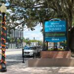 On the left is a light pole with autumn-colored leaves hanging from it. To the right of that is a sign that reads, "Historic Downtown Square Ocala." In the background is a large tree, shading both the sign and the light pole. Further behind the tree is a white gazebo on the right and slow traffic on the left.