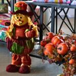 A smiling scarecrow dressed in fall colors stands next to a shelf with fake pumpkins, leaves and vine. Behind the scarecrow are quilts of varying colors.