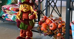 A smiling scarecrow dressed in fall colors stands next to a shelf with fake pumpkins, leaves and vine. Behind the scarecrow are quilts of varying colors.