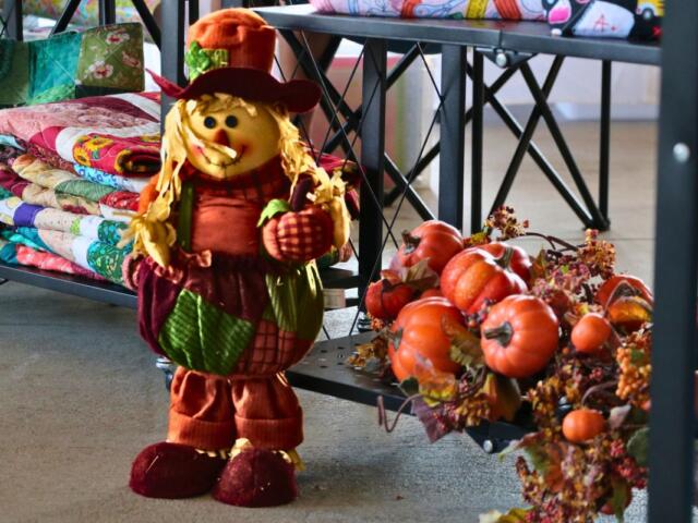 A smiling scarecrow dressed in fall colors stands next to a shelf with fake pumpkins, leaves and vine. Behind the scarecrow are quilts of varying colors.
