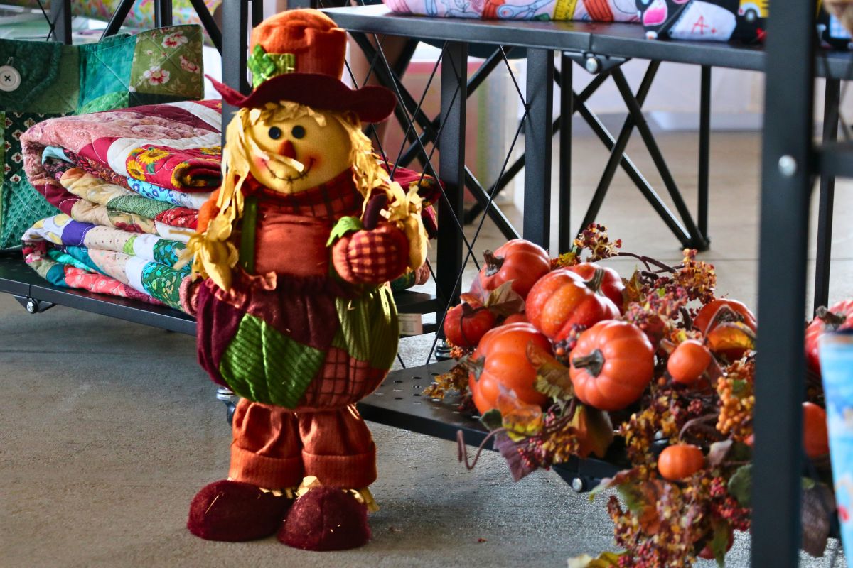 A smiling scarecrow dressed in fall colors stands next to a shelf with fake pumpkins, leaves and vine. Behind the scarecrow are quilts of varying colors.
