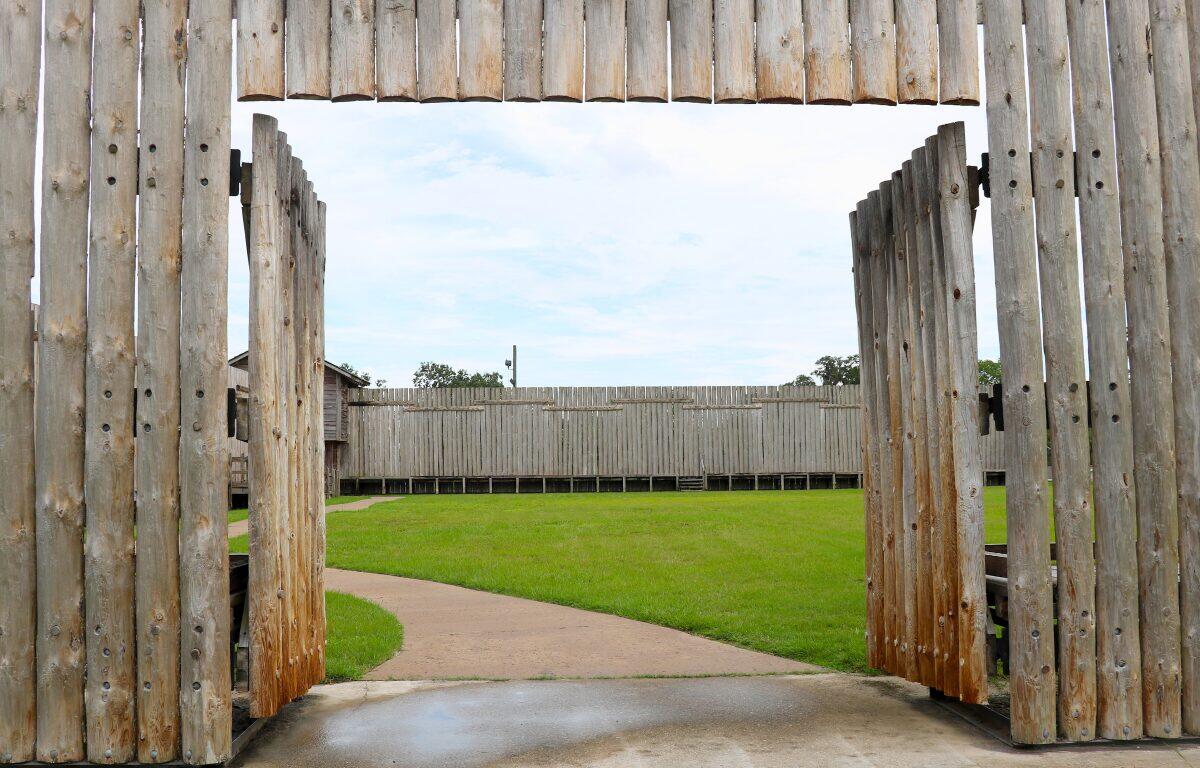 A tall wooden wall and open double door host an open grass space with a path leading to another wooden wall perpendicular to the one on the foreground.