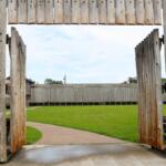 A tall wooden wall and open double door host an open grass space with a path leading to another wooden wall perpendicular to the one on the foreground.