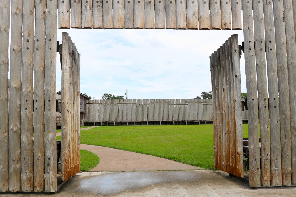 A tall wooden wall and open double door host an open grass space with a path leading to another wooden wall perpendicular to the one on the foreground.