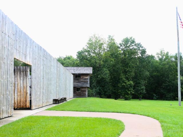 There is an open grass field with trees in the distance. On the right lies a tall fall pole with the American flag flying at the top. To left is a large wooden wall with open doors and a tower at the corner.