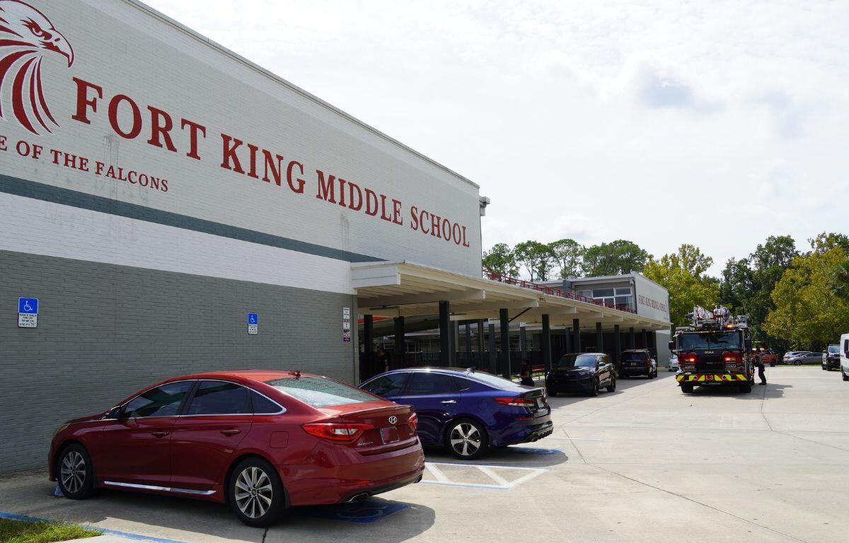 A building with a white facade, with writing on it in the background, with cars parked in a lot in front of the building. To the far right, there's a platform shelter with cars in front of it.