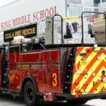 A red fire truck in front of a structure with writing on the facade, and a widow on the right of the structure.