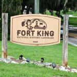 A beige sign held up on either side with wood pillars and lights below reads, "FORT KING NATIONAL HISTORIC LANDMARK 3925." On the sign is a drawing of a fort and s Seminole. In the background is a road with a car and trees.
